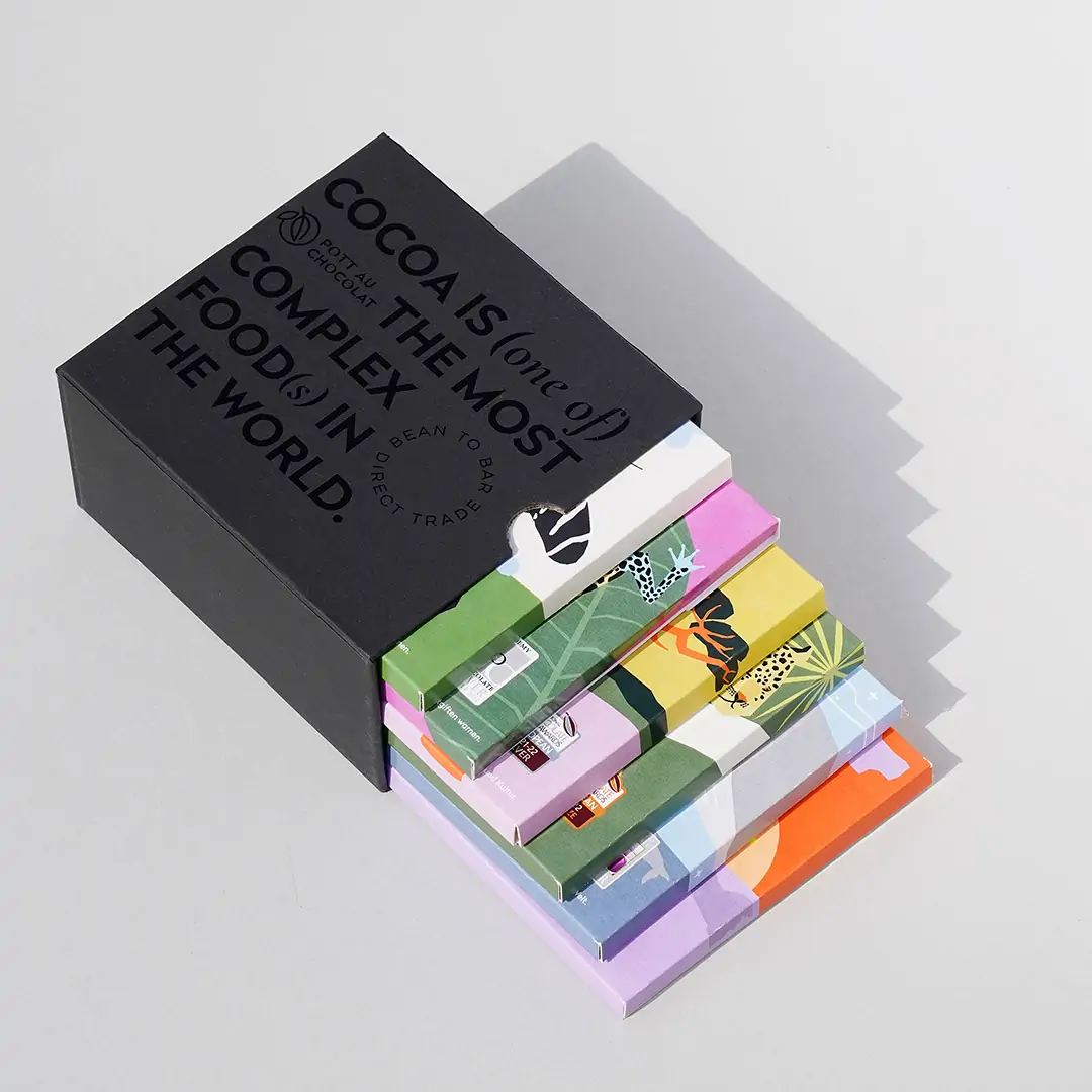 6 bunte Schokoladentafeln in einem schwarzen Schuber aus Pappe, der mit schwarzer Schrift geprägt wurde. Der Text ist englisch und der Inhalt: Kakao ist eines der komplexesten Lebensmittel der Welt. Die Schokoladentafeln sind so aus der Box gezogen, dass man jede etwas sehen kann.
