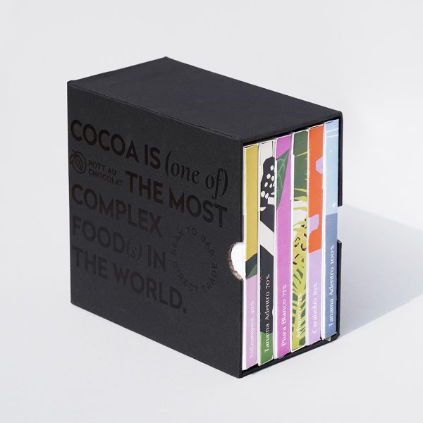 Unsere Mix and Match Chocolate Gift Box - 6 bunte Schokoladentafeln verpackt in einem schwarzen Schuber aus Pappe, der mit schwarzer Schrift geprägt wurde. Der Text ist englisch und der Inhalt: Kakao ist eines der komplexesten Lebensmittel der Welt.