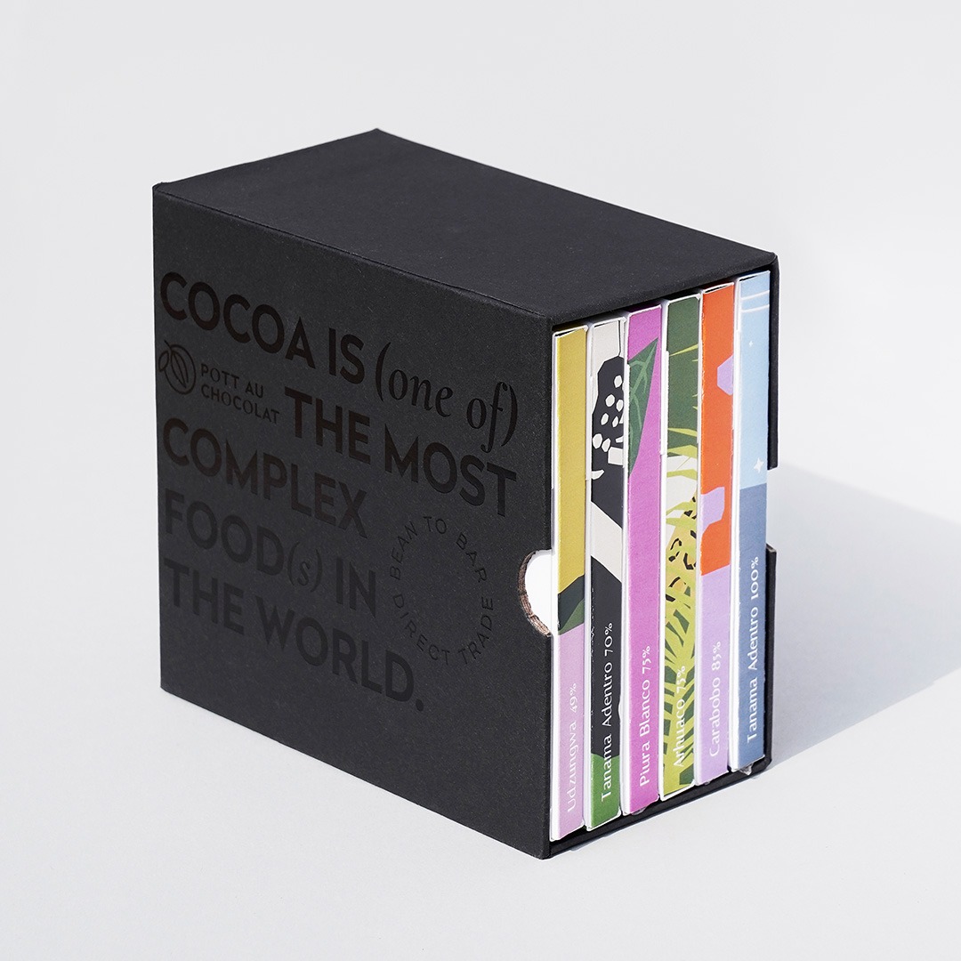 Unsere Mix and Match Chocolate Gift Box - 6 bunte Schokoladentafeln verpackt in einem schwarzen Schuber aus Pappe, der mit schwarzer Schrift geprägt wurde. Der Text ist englisch und der Inhalt: Kakao ist eines der komplexesten Lebensmittel der Welt.