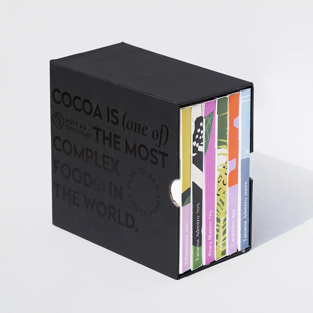 Unsere Mix and Match Chocolate Gift Box - 6 bunte Schokoladentafeln verpackt in einem schwarzen Schuber aus Pappe, der mit schwarzer Schrift geprägt wurde. Der Text ist englisch und der Inhalt: Kakao ist eines der komplexesten Lebensmittel der Welt.