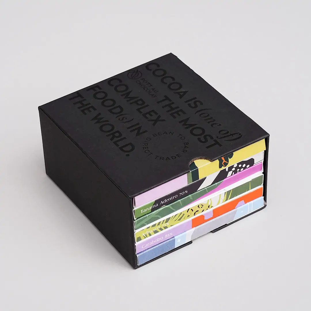 Schwarzer Schuber aus Pappe, der mit schwarzer Schrift geprägt wurde. Der Text ist englisch und der Inhalt: Kakao ist eines der komplexesten Lebensmittel der Welt. Darin sieht man 6 bunte Schokoladentafeln.