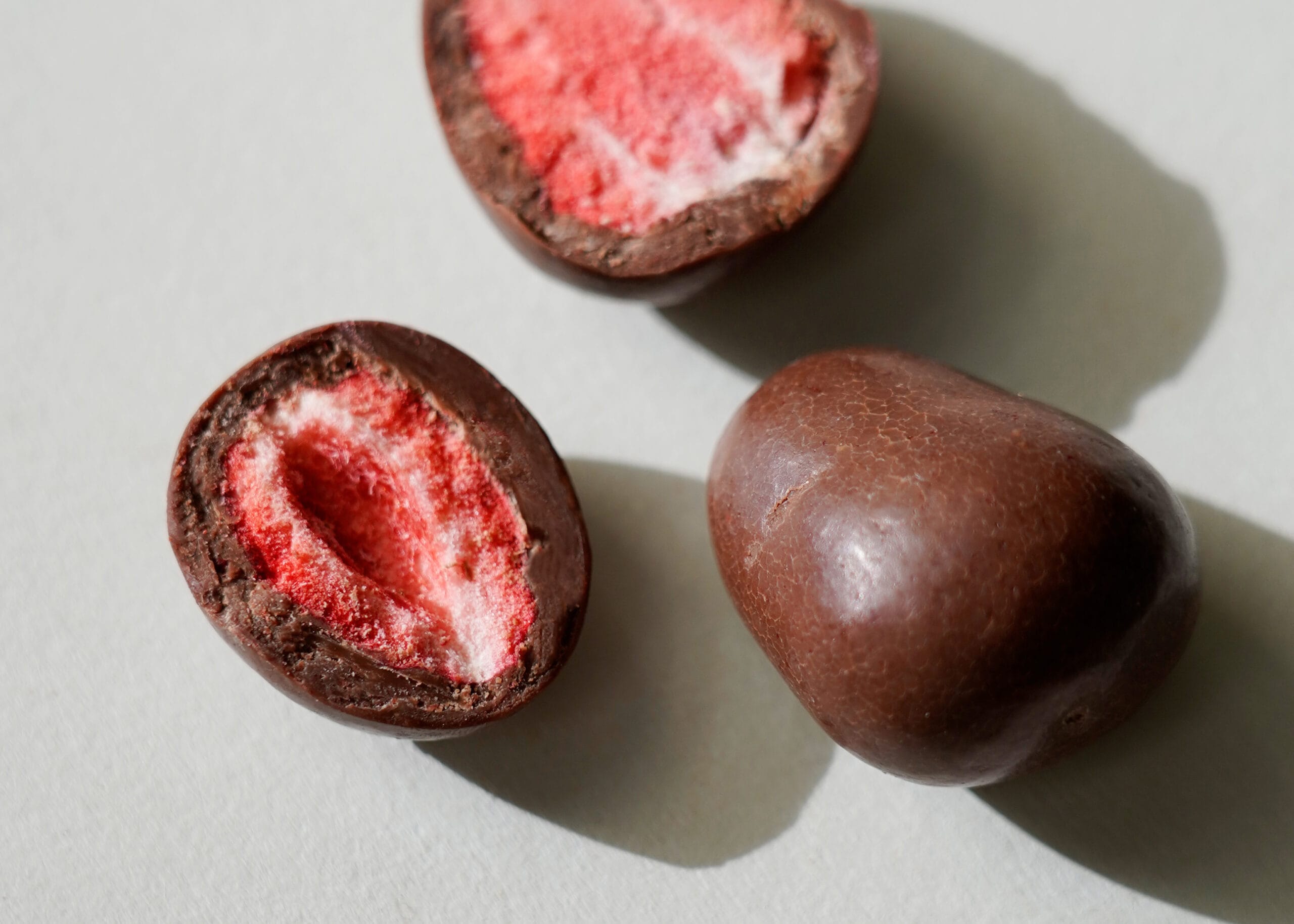 Nahaufnahme von dragierten gefriergetrockneten Erdbeeren. Eine ganz, die anderen beiden jeweils zur Hälfte aufgeschnitten, sodass man die rote Erdbeere im Inneren und die Stärke der Milchschokoladenhülle erkennen kann.