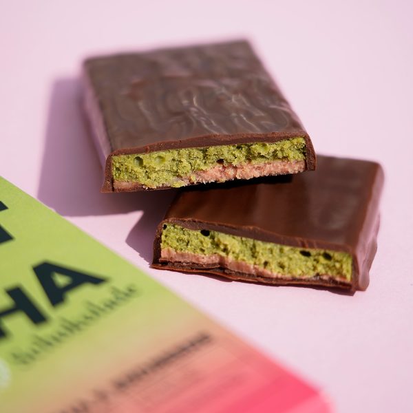 Mit dunkler Schokolade überzogene gefüllte Tafel, aufgeschnitten. Die Füllung besteht aus einer rosafarbenen und grünen Schicht. Daneben liegt unscharf die Verpackung.