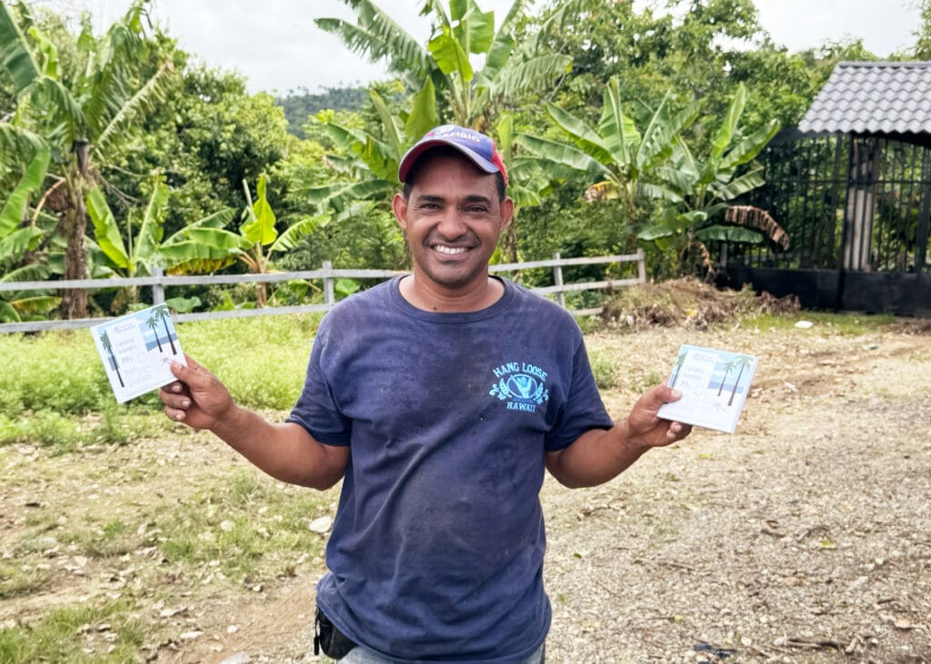 Arcenio steht vor einer Kakao Plantage und hält zwei unserer Tanama 39% Tafeln in die Kamera.