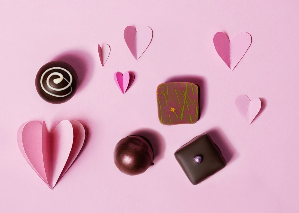 Auf einem rosa Hintergrund liegen rosa Herzen aus Papier und 4 Pralinen.