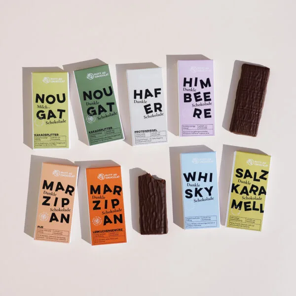 Mehrere Verpackungen der gefüllten Tafeln sind zu sehen – Marzipan Lebkuchengewürz, Marzipan Pur, Whisky, Salzkaramell, Himbeere, Nougat Kakaosplitter mit Milchschokolade, Nougat Kakaosplitter mit dunkler Schokolade sowie Hafer. Die Verpackungen sind bunt gestaltet in Orange, Hellblau, Gelb, Rosa, Dunkelgrün, Hellgrün, Beige und Hellorange. Zwischen den Verpackungen liegen zwei Tafeln der gefüllten Schokolade