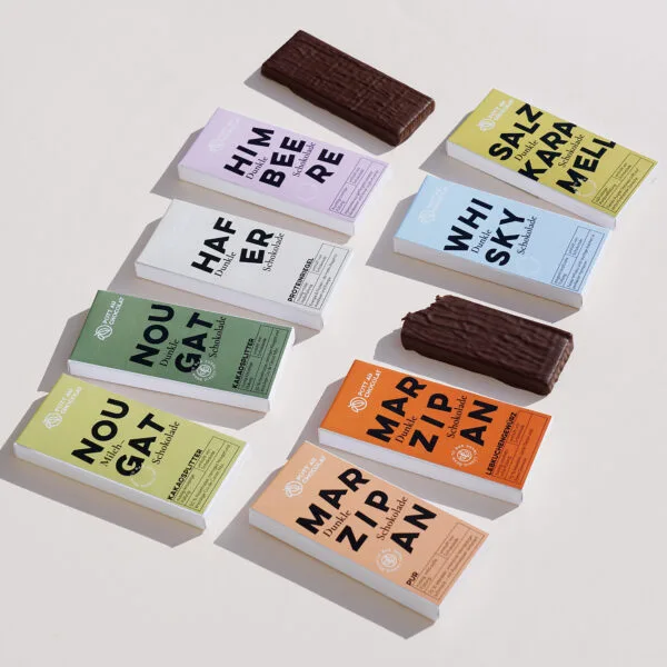 Mehrere Verpackungen der gefüllten Tafeln sind zu sehen – Marzipan Lebkuchengewürz, Marzipan Pur, Whisky, Salzkaramell, Himbeere, Nougat Kakaosplitter mit Milchschokolade, Nougat Kakaosplitter mit dunkler Schokolade sowie Hafer. Die Verpackungen sind bunt gestaltet in Orange, Hellblau, Gelb, Rosa, Dunkelgrün, Hellgrün, Beige und Hellorange. Zwischen den Verpackungen liegen zwei Tafeln der gefüllten Schokolade