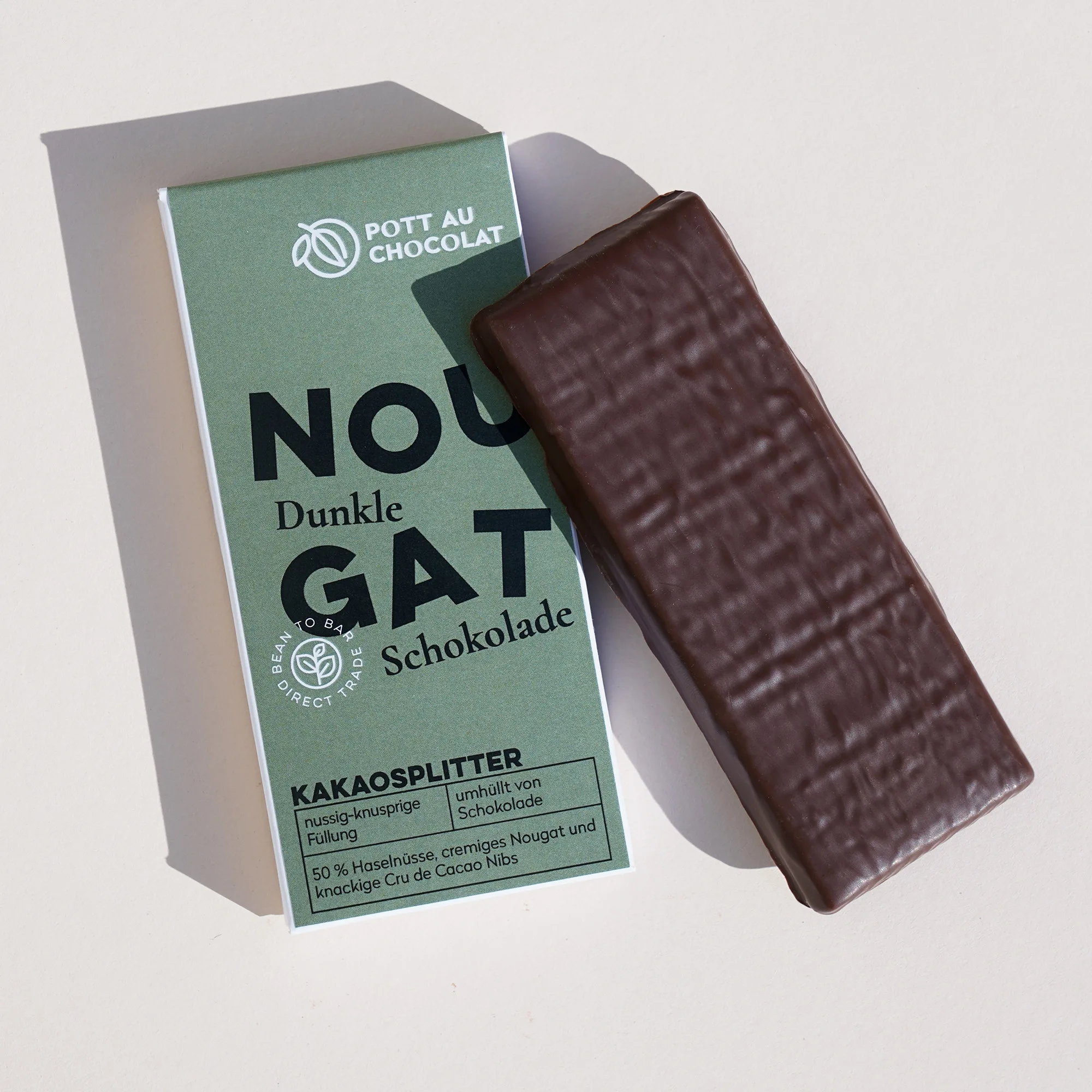 Gefüllte Tafel aus dunkler Schokolade. Daneben die Verpackung mit der Aufschrift 'Nougat Kakaosplitter'. Die Aufschrift ist verspielt mit tanzenden Buchstaben gestaltet. Farben des Designs: Dunkles Grün, Schwarz und Weiß