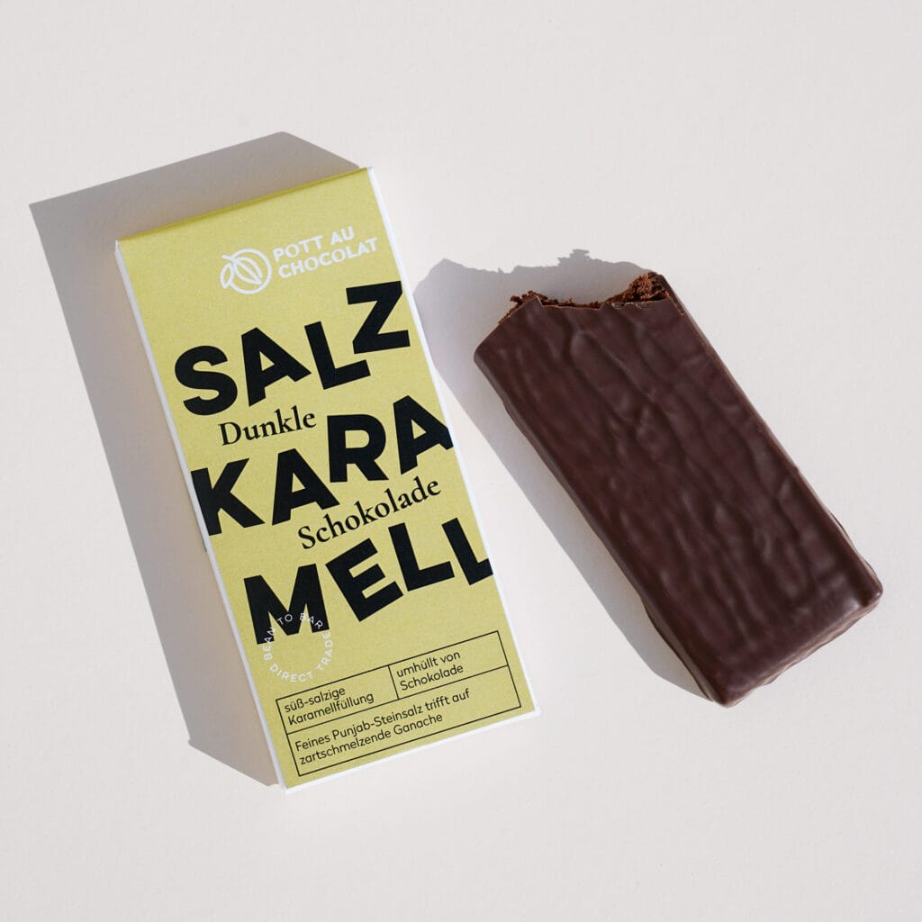 Gefüllte Tafel aus dunkler Schokolade. Daneben die Verpackung mit der Aufschrift 'Salzkaramell'. Die Aufschrift ist verspielt mit tanzenden Buchstaben gestaltet. Farben des Designs: Gelb, Schwarz und Weiß