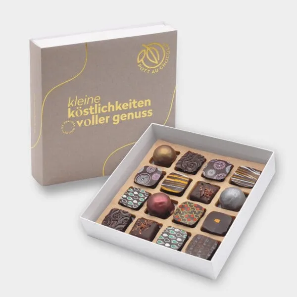 Aufgestellte schlichte Pralinenbox in Grau mit goldgeprägter Schrift: 'Kleine Köstlichkeiten voller Genuss'. Davor eine offene Box mit 16 bunt gemischten Pralinen