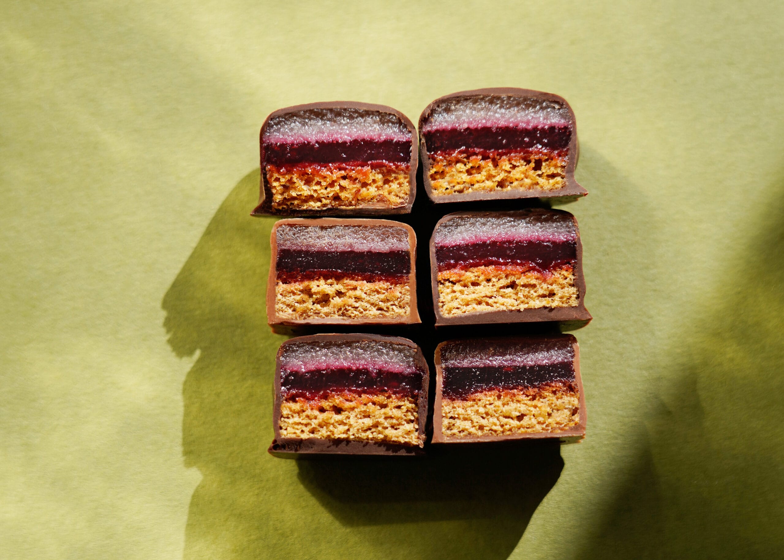 Pott-au-Chocolat-Dominosteine-angeschnitten-9 zwei Reihen von je drei zur Hälfte angeschnittenen Dominosteinen von Pott au Chocolat vor grünem Hintergrund zeigen den Schichtaufbau: Honigkuchenteil, Fruchtgelee Himbeere-Johannisbeere, Marzipan und alles umhüllt von Schokolade