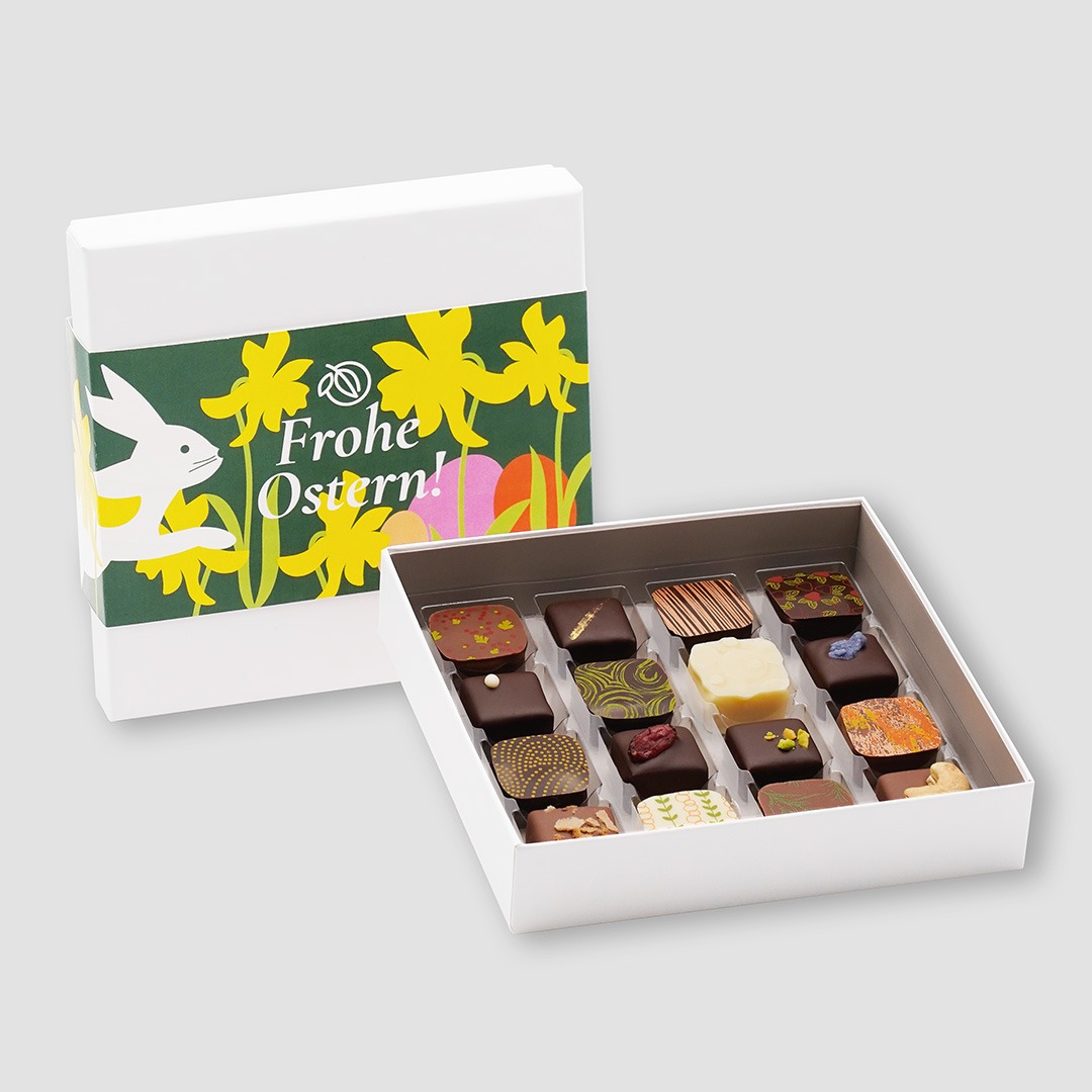 Weiße Pralinenbox mit 16 bunt dekorierten Pralinen. Dahinter steht die geschlossene Schachtel mit einer Banderole in dunkelgrün mit 'Frohe Ostern' und mit gelben Narzissen, Hase und bunten Eiern.