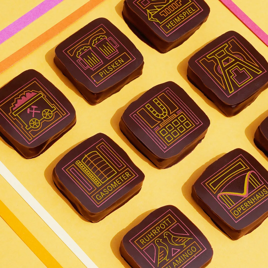 9 Pralinen in dunkler Schokolade auf gelbem Hintergrund. Die Pralinen sind mit Motiven aus Kakaobutter bedruckt, die verschiedene Ruhrgebiets Bilder zeigen. Dortmunder U, Gasometer, Stadion, Förderturm und weitere. In den Farben gelb, orange und pink.