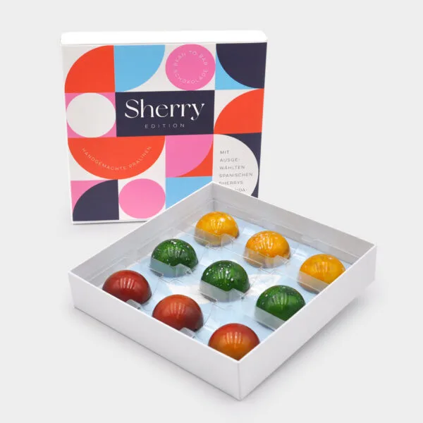 Aufgestellte Pralinenbox mit buntem Design in geometrischen Formen und der Aufschrift 'Sherry Edition'. Davor die geöffnete Box mit den Sherry-Pralinen. Farben des Designs: Hellblau, Hellrot, Weiß, Rosa, Dunkles Lila