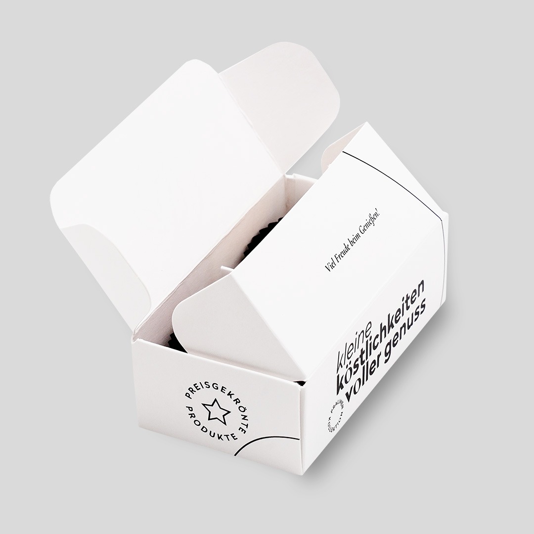 Halb geöffnete Verpackung für zwei Macarons aus weißem Karton, bedruckt mit schwarzer Schrift 'Kleine Köstlichkeiten voller Genuss' und dem Logo von Pott au Chocolat.