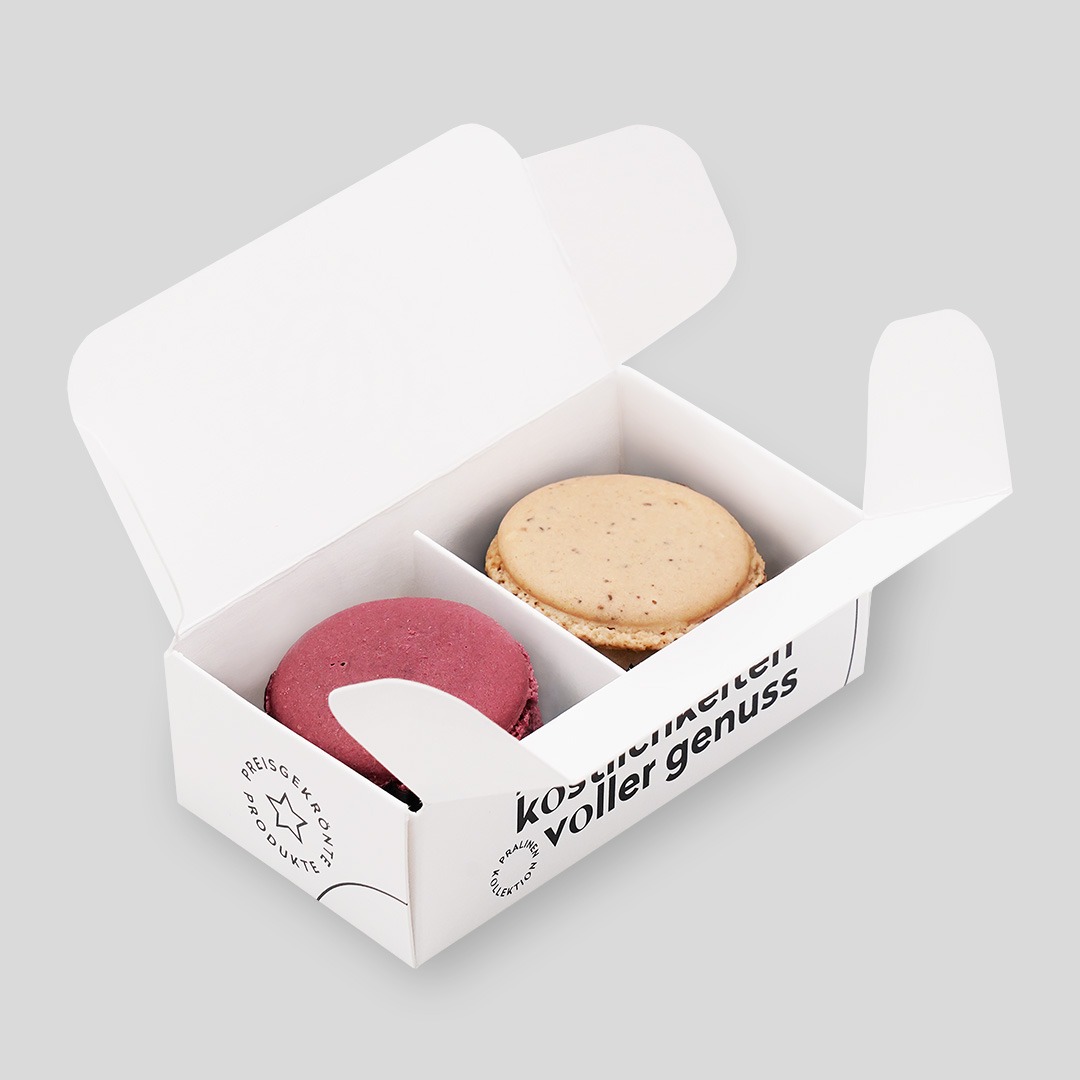Kleine Box aus weißem Karton mit schwarzer Schrift. Geöffnet, darin sind zwei Macarons, in Pink und Beige
