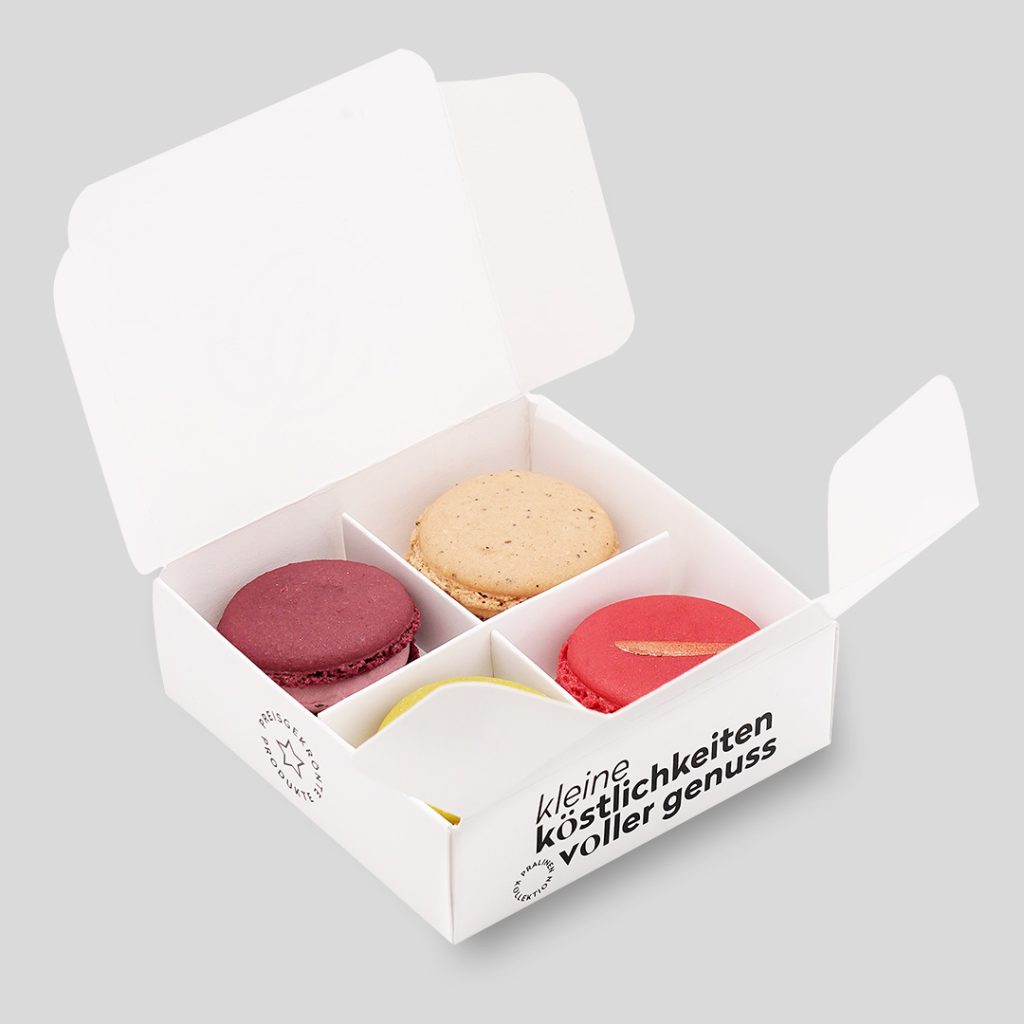 Kleine geöffnete Box, aus weißem Karton mit schwarzer Schrift: 'Kleine Köstlichkeiten voller Genuss', 'Preisgekrönte Produkte'. Darin sind vier Macarons, in Pink, Beige, Grün und Beere