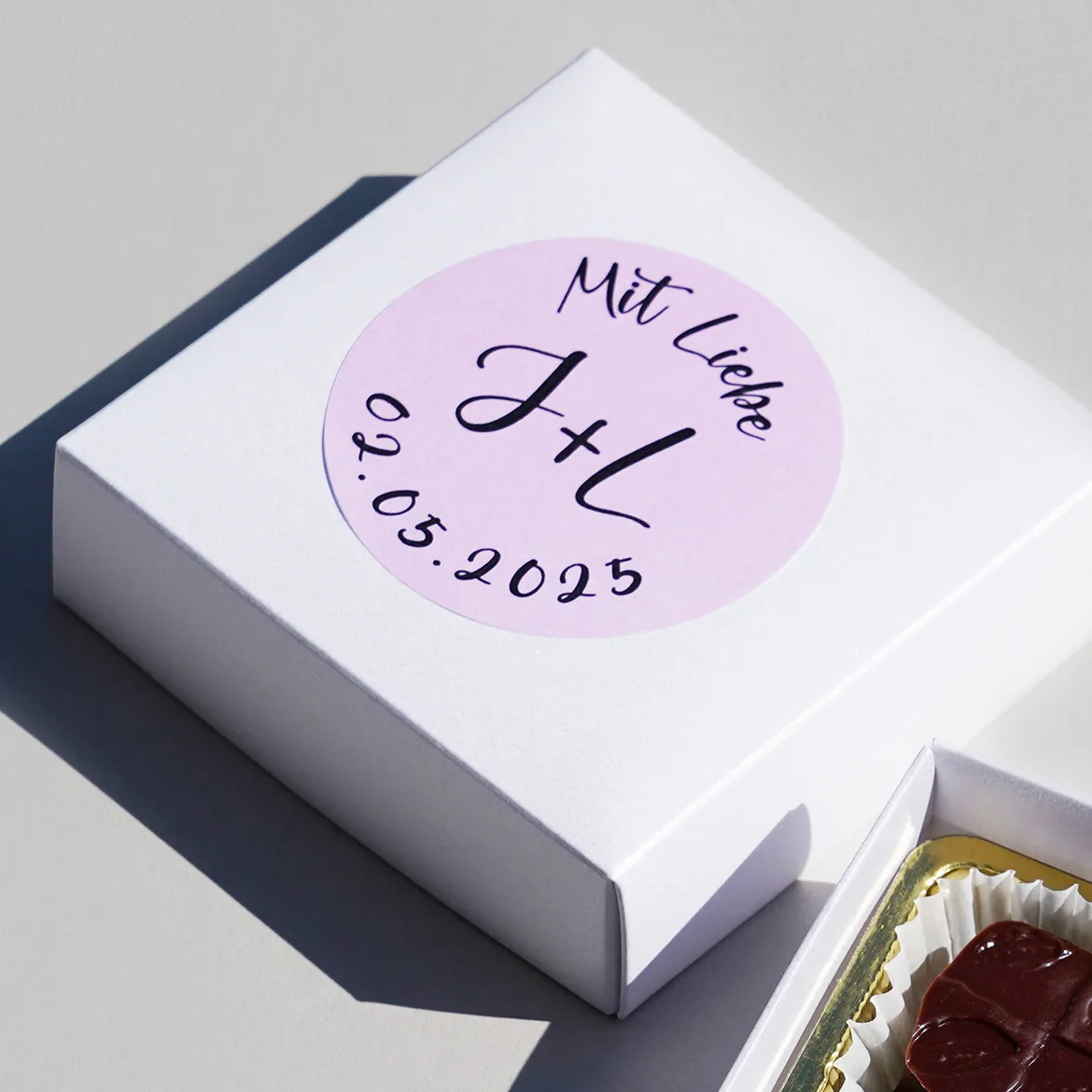 Close-Up von 4 Pralinen in einer kleinen weissen, geöffneten Geschenkbox daneben liegt ein Beispielhaft für ein Hochzeits-Give-Away mit einem runden Etikett individualisierter Deckel