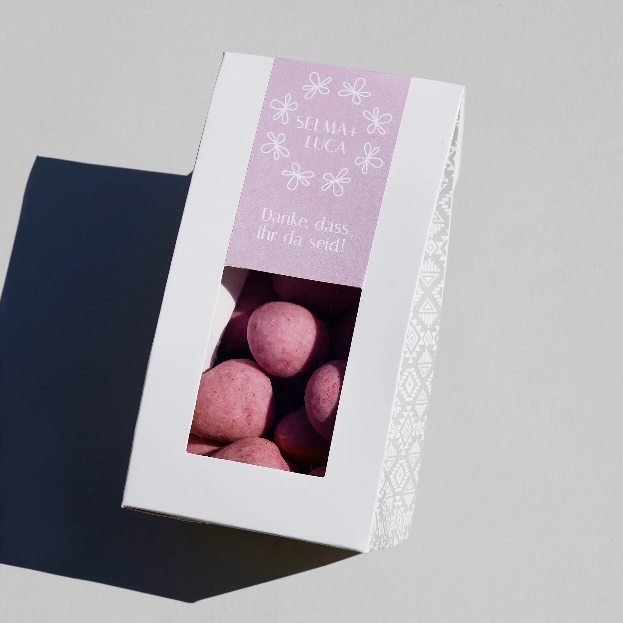 Gibelverpackung mit Fenster versehen mit einem individualisierten Etikett als Beispiel für eine Platzkarte auf einem Hochzeitstisch. Die Verpackung ist gefüllt mit unseren Dragierten Himbeeren in weisser Schokolade vermischt mit Himbeerstaub