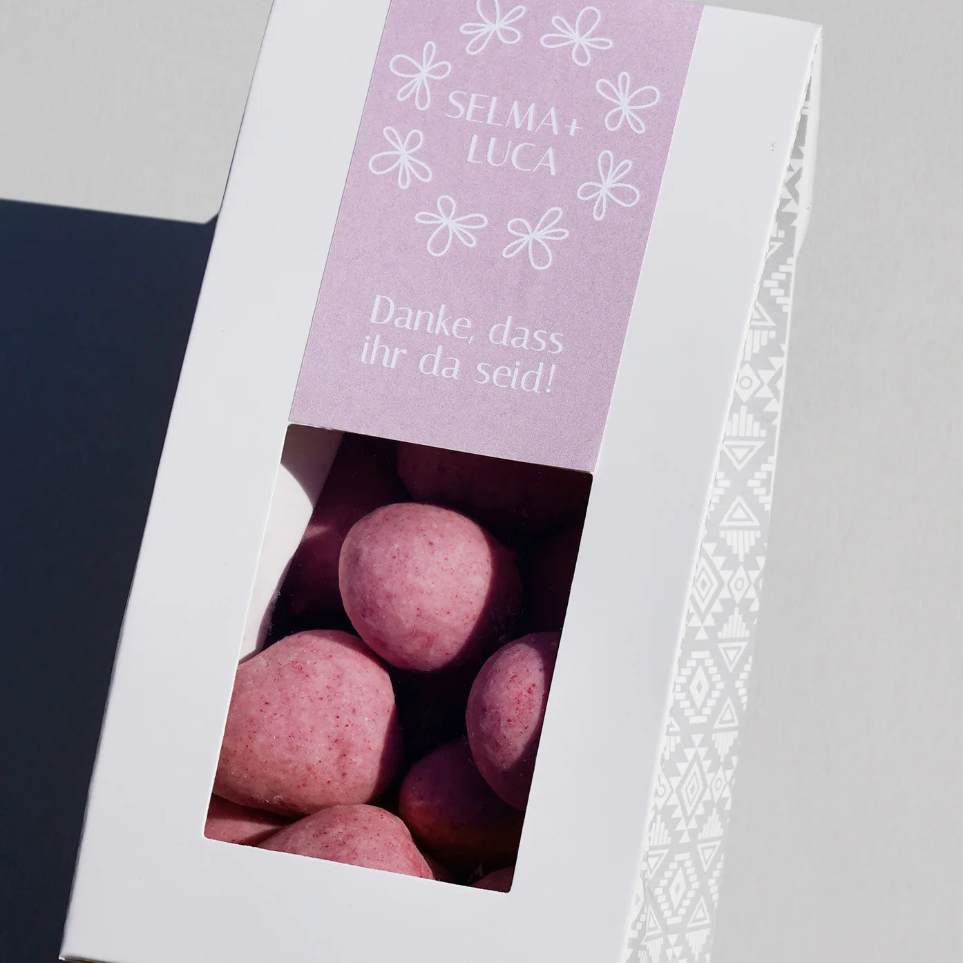 Gibelverpackung mit Fenster versehen mit einem individualisierten Etikett als Beispiel für eine Platzkarte auf einem Hochzeitstisch. Die Verpackung ist gefüllt mit unseren Dragierten Himbeeren in weisser Schokolade vermischt mit Himbeerstaub
