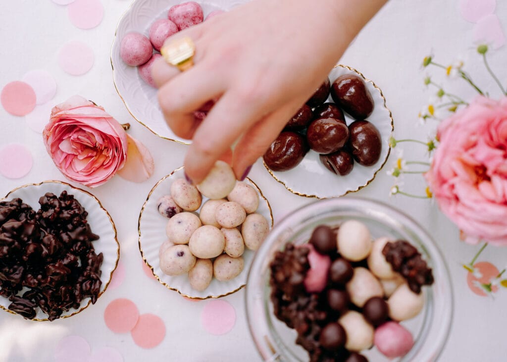 Eine Hand greift eine Amarenakirsche in Schokolade von einem Buffet. Es stehen mehrere Glasschalen mit dragierten Produkten auf einem Tisch. Dazu stehen da Blumen und es liegt Konfetti.