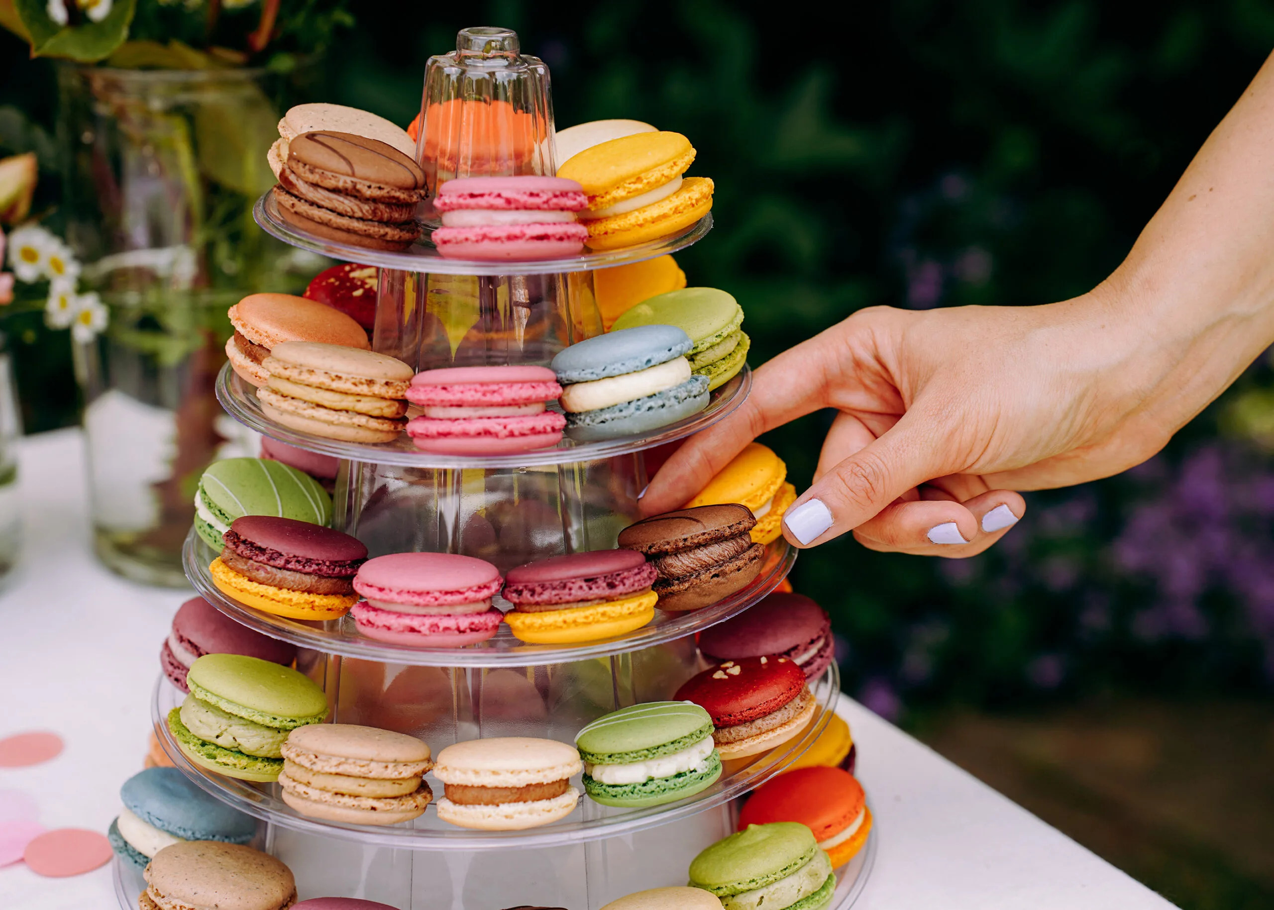 Eine Hand greift einen Mararon auf einer Macaron Pyramide, auf der viele bunte Macarons liegen. Es ist ein Buffet im Garten auf einer Hochzeit.