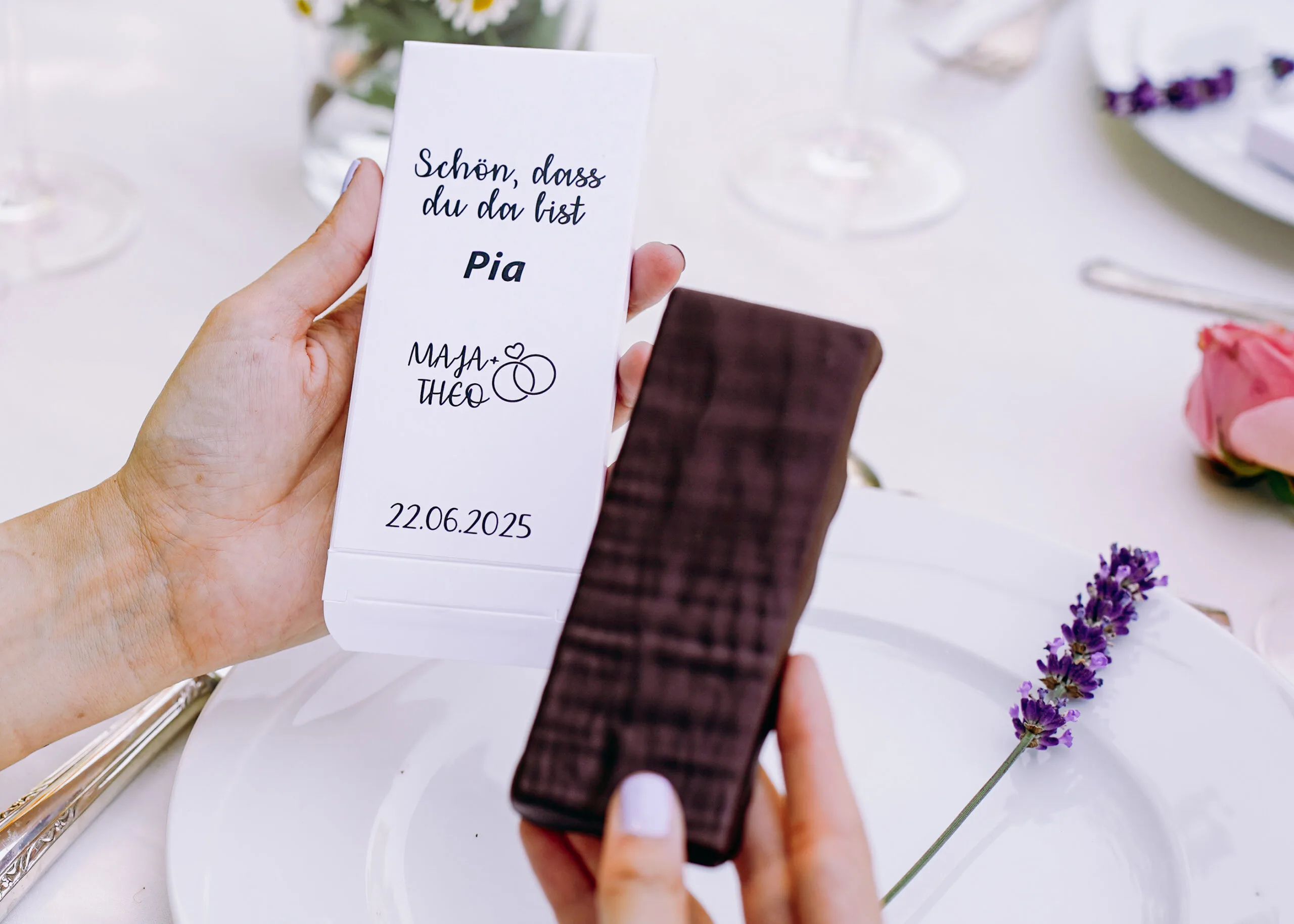 Eine Hand hält eine Schokoladentafel, in der anderen die Verpackung mit der Aufschrift 'Schön, dass du da bist', einem Namen, den Namen eines Brautpaares und das Hochzeitsdatum. Die Person sitzt an ihrem Platz an der Hochzeitstafel.