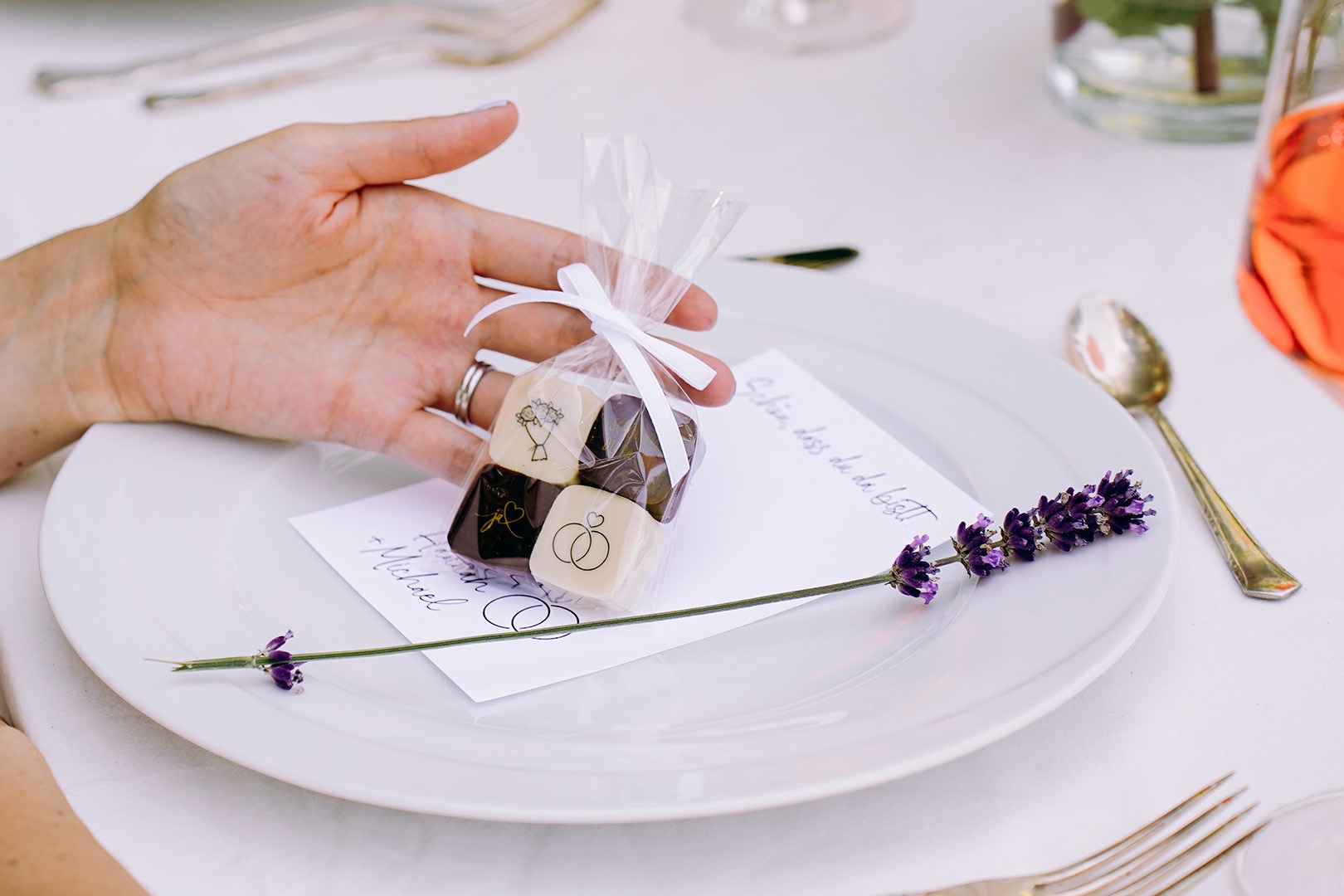 Eine Hand hält ein Tütchen mit 4 Pralinen mit Hochzeitsmotiven an einem festlich gedeckten Tisch