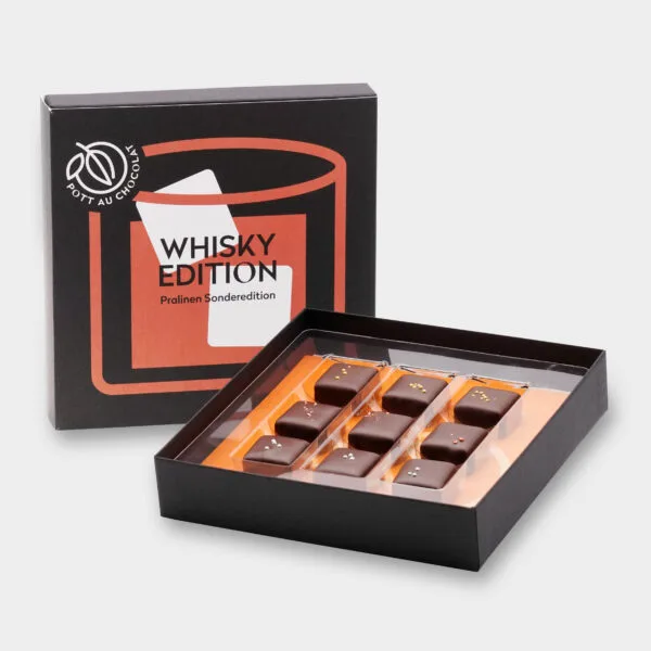 Aufgestellte Pralinenbox 'Whisky Edition'. Design in den Farben Schwarz, Orange und Weiß mit einer abstrakten Darstellung eines Glases mit Whisky und Eiswürfeln. Davor die geöffnete Box mit den Whisky-Pralinen