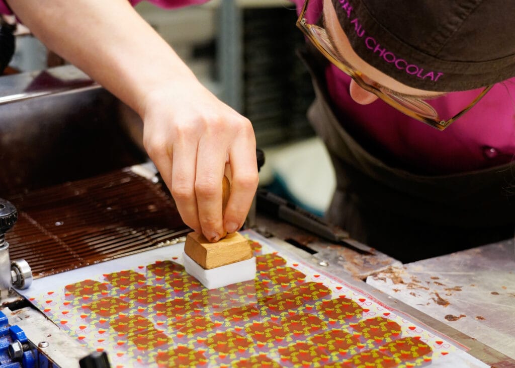 Nach dem Überzug erhalten die Schnitt-Pralinen ihr Dekor. Hier in Form einer Transferfolie, die eine Mitarbeiterin gerade auf einige Pralinen aufgelegt hat und jetzt mit einem Stempel auf die Pralinen drückt.