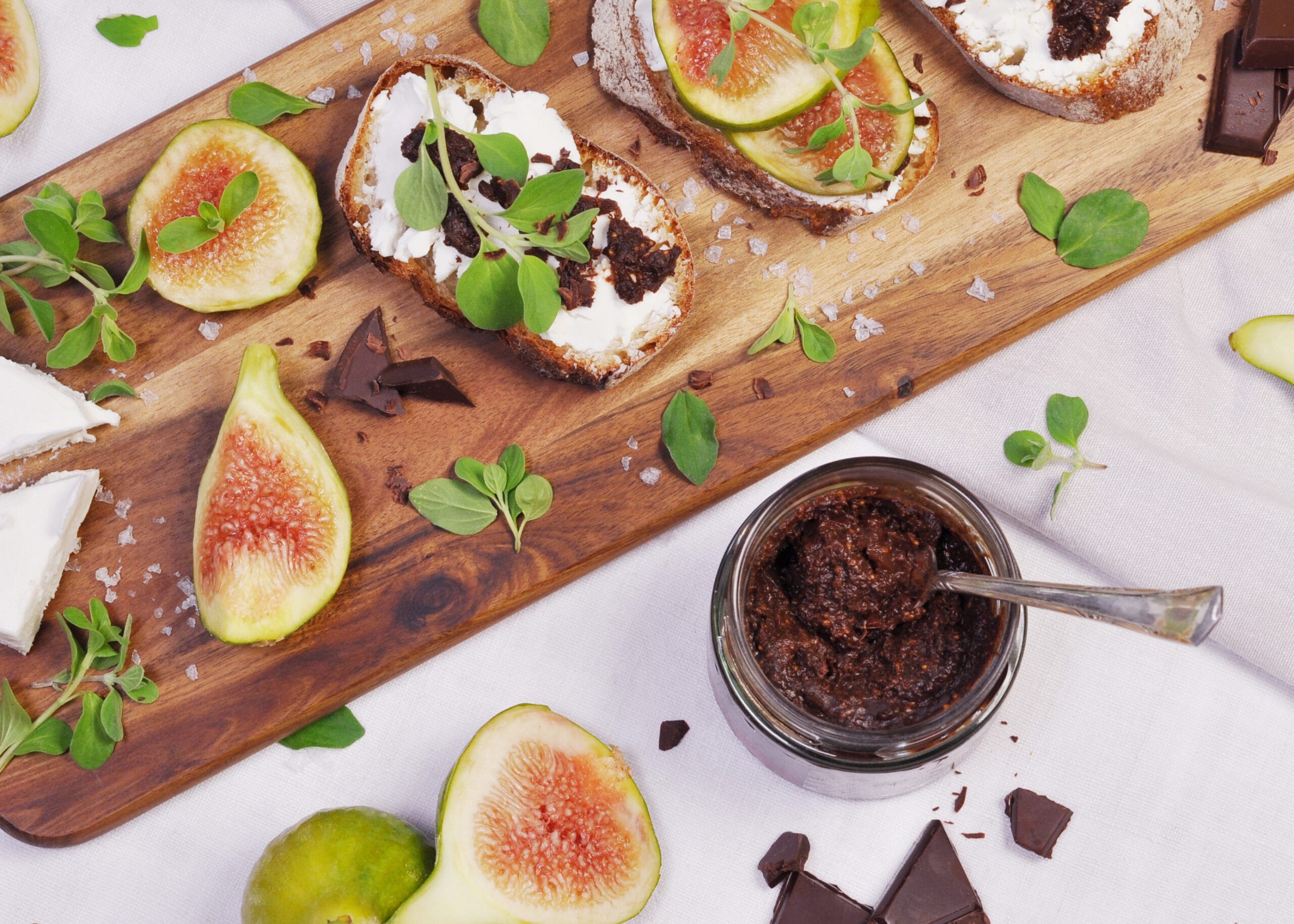 angerichteter Tisch von oben fotografiert mit einem Holzbrett. Darauf aufgeschnittene Feigen und Brote mit Käse und etwas Feigen Schokoladen Chutney, das auch in einem geöffneten Glas mit Löffel im vorderen Bereich des Fotos zu sehen ist.