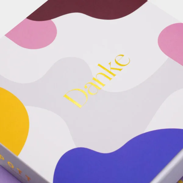 Angeschnittene Nahaufnahme Pralinenbox "Danke" schräg von oben: bunte organischen Formen mit der goldgeprägten Aufschrift 'Danke'.