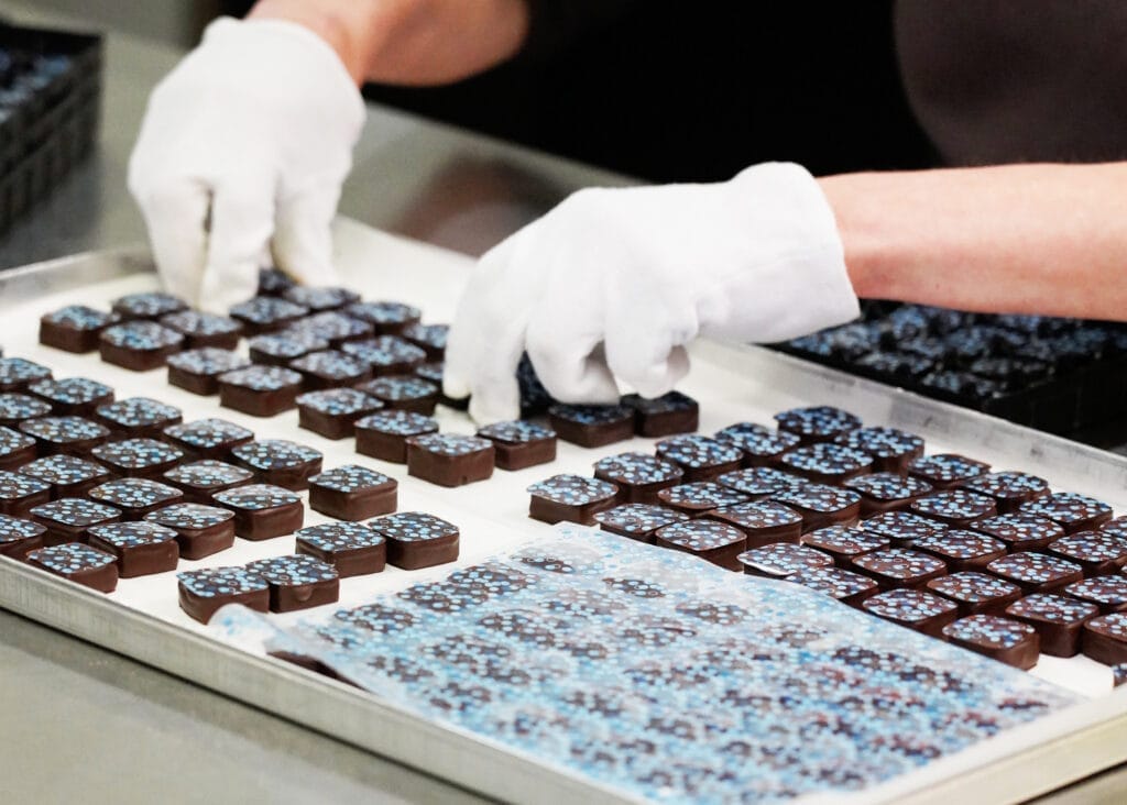 Viele Heidelbeer Pralinen mit blauem Dekor auf einem Backblech, die von einer Mitarbeiterin mit Handschuhen vom Blech genommen werden. Im Vordergrund eine Charge Pralinen unter der Transferfolie