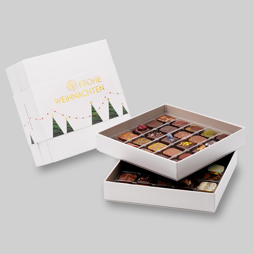 Pralinenbox Weihnachten 50 Pralinen - mit einer geöffneten Schachtel mit zwei Lagen, mit jeweils 25 bunt dekorierten Pralinen. Die Verpackung ist in weiß gehalten mit goldener Schrift 'Frohe Weihnachten' und grünen Bäumchen.