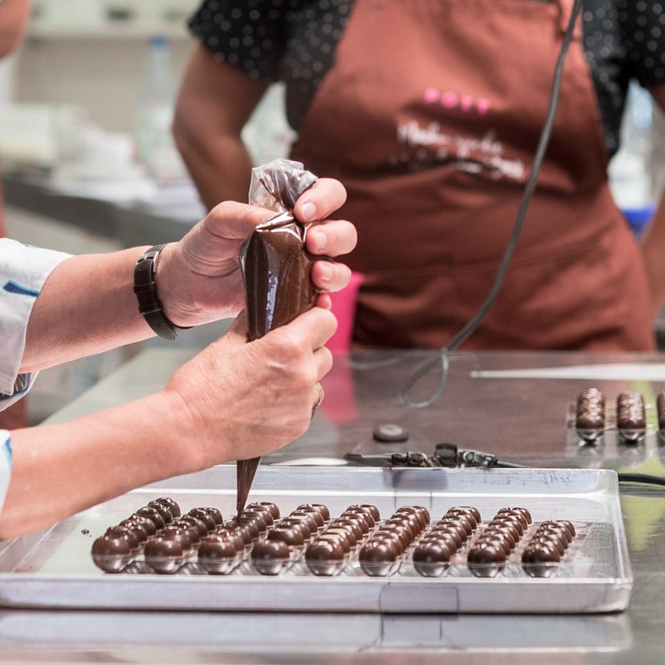 Ein Pralinenseminar Teilnehmer beim spritzen von einer Pralinenfüllung (Ganache) in Hohlkörper. Im Hintergrund ein zweiter Teilnehmer beim zusehen.