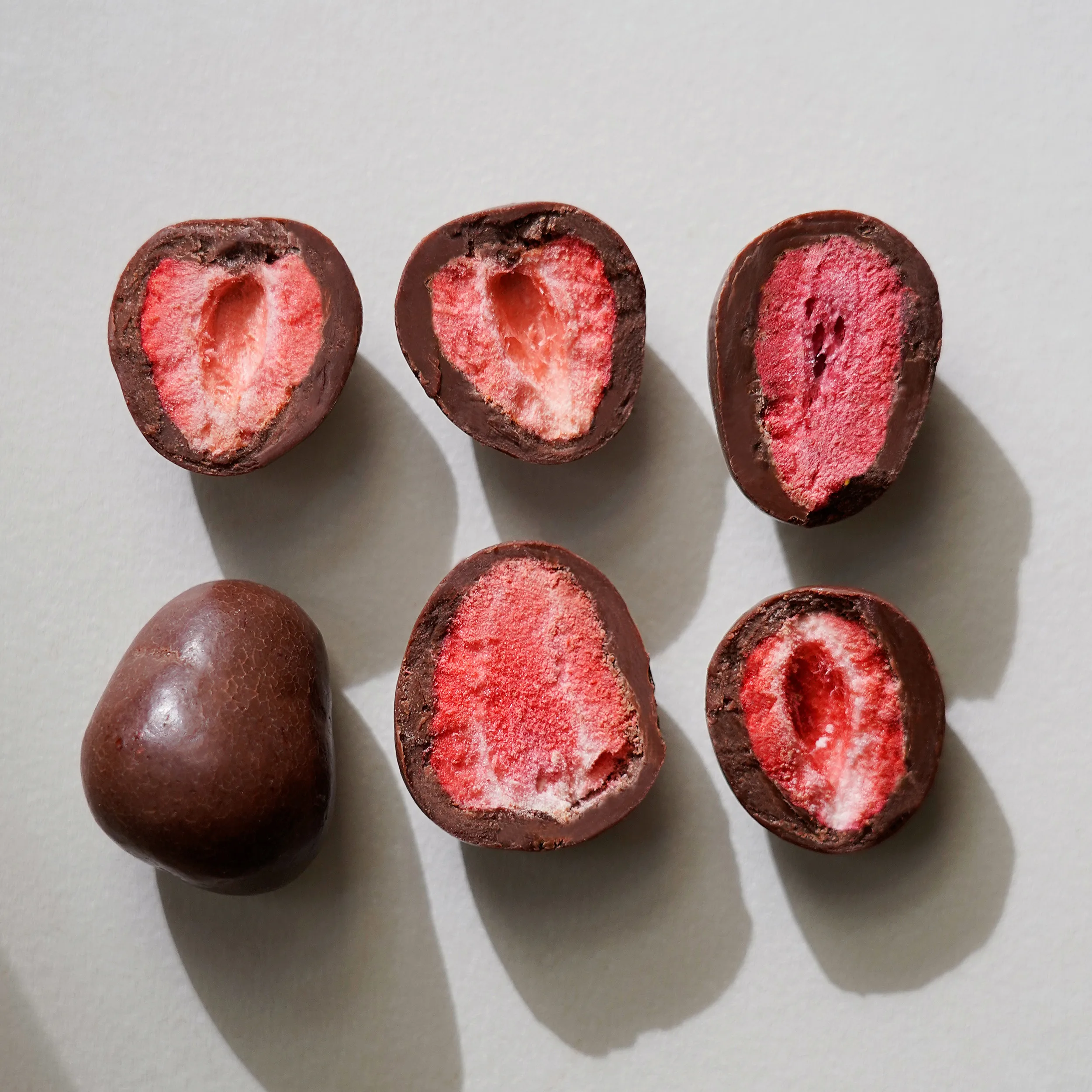 Zwei Reihen á 3 Stück von dragierten gefriergetrockneten Erdbeeren. Fünf zur Hälfte eingeschnitten und links unten eine ganze schokolierte Frucht.