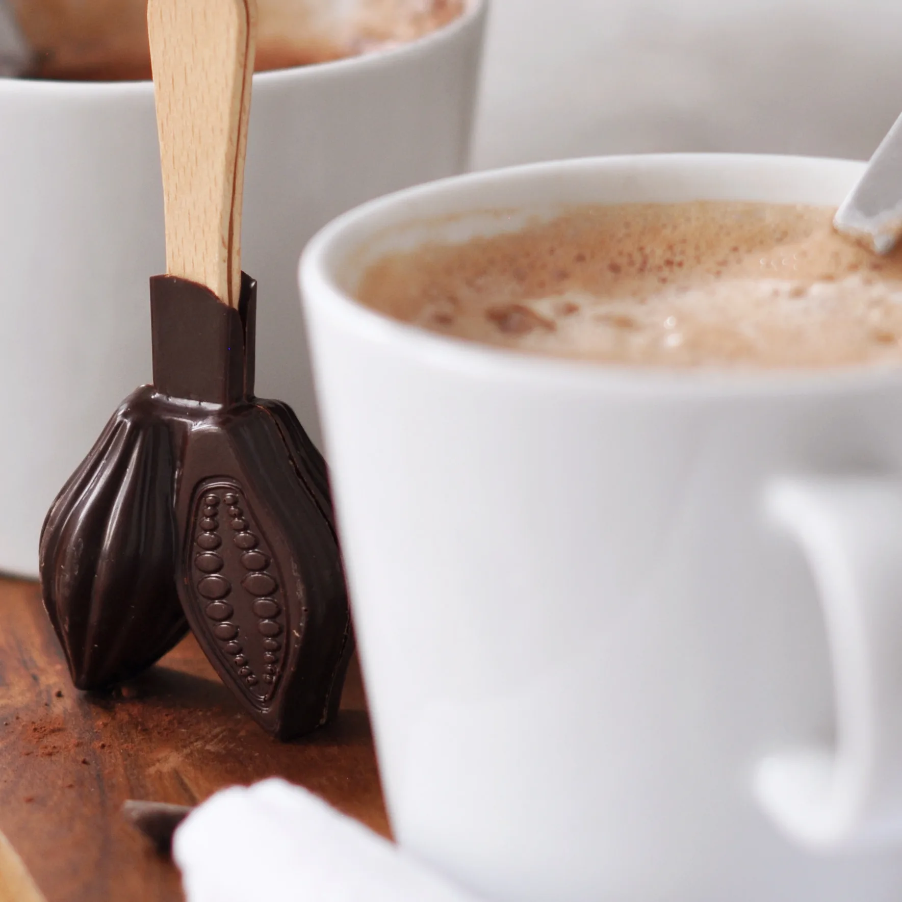 Nahaufnahme einer weißen Tasse mit schaumig gerührter Trinkschokolade dahinter unser dunkler Schokoladenstick Heiß am Stiel