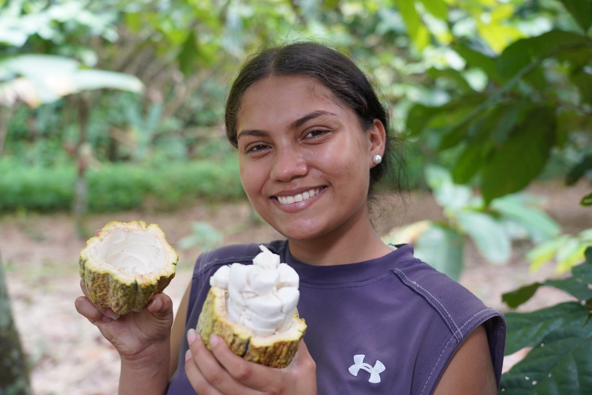 Eine lachende junge Venezolanerin hält eine aufgebrochene gelbe Kakaofrucht in die Kamara. Im Hintergrund stehen Kakaobäume. Aufgenommen von Silva Cacao in der Region Puerto Cabello in Venezuela