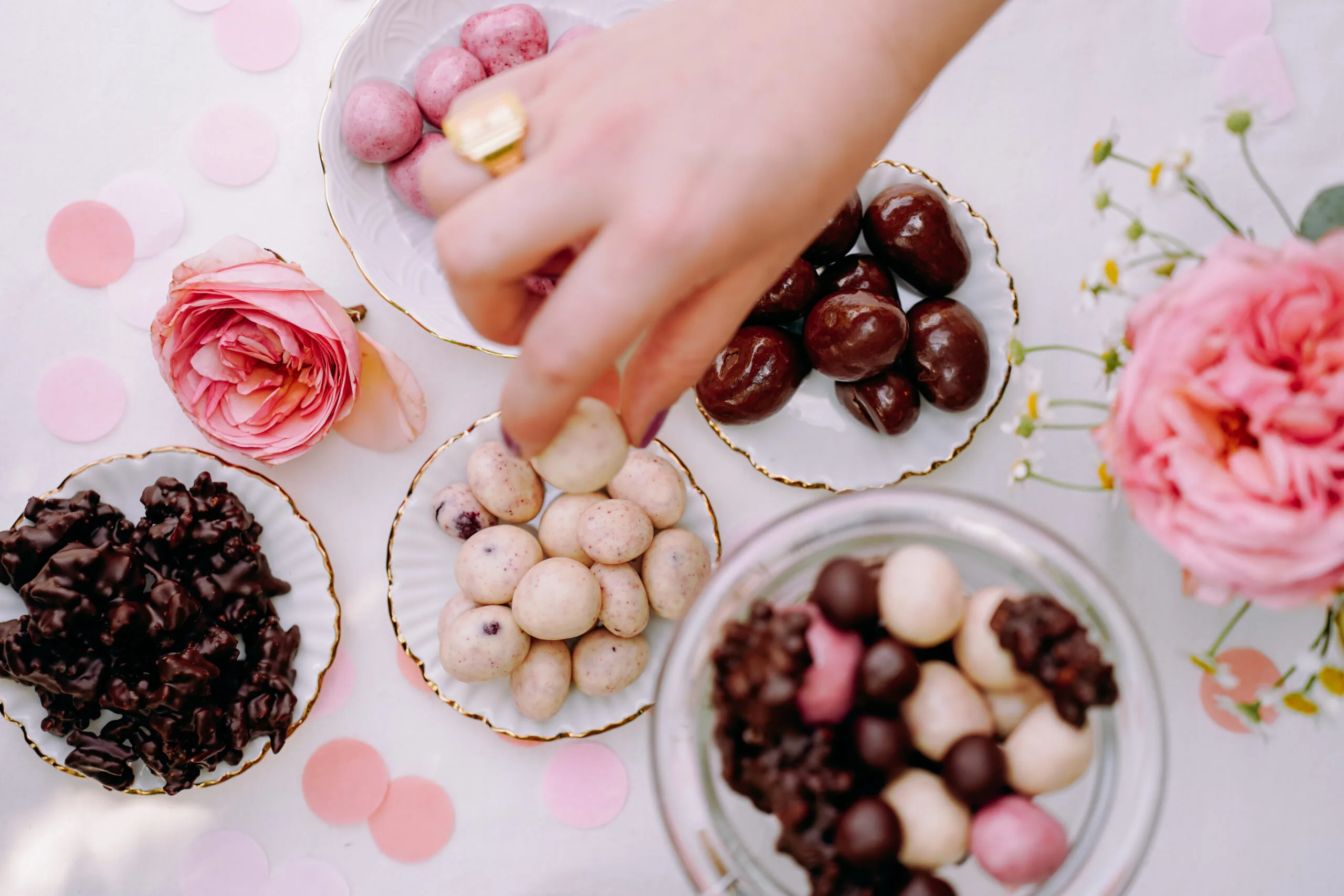 Eine Hand greift eine Amarenakirsche in Schokolade von einem Buffet. Es stehen mehrere Glasschalen mit dragierten Produkten auf einem Tisch. Dazu stehen da Blumen und es liegt Konfetti.