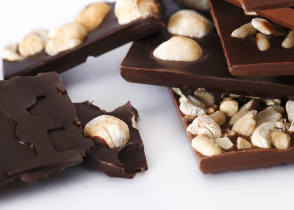 Gebrochene Schokoladentafeln mit verschiedenen Nüssen belegt auf einem Haufen. Macadamia und Cashew